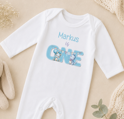 A white onesie with the text "Markus is ONE" printed on it, along with illustrations of a cat and a dog.