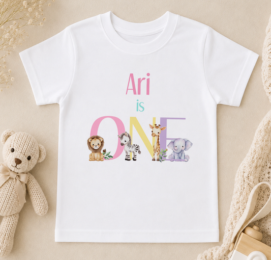 A white t-shirt with the text "Ari is ONE" printed on it, featuring various animal illustrations such as a lion, zebra, giraffe, and elephant.