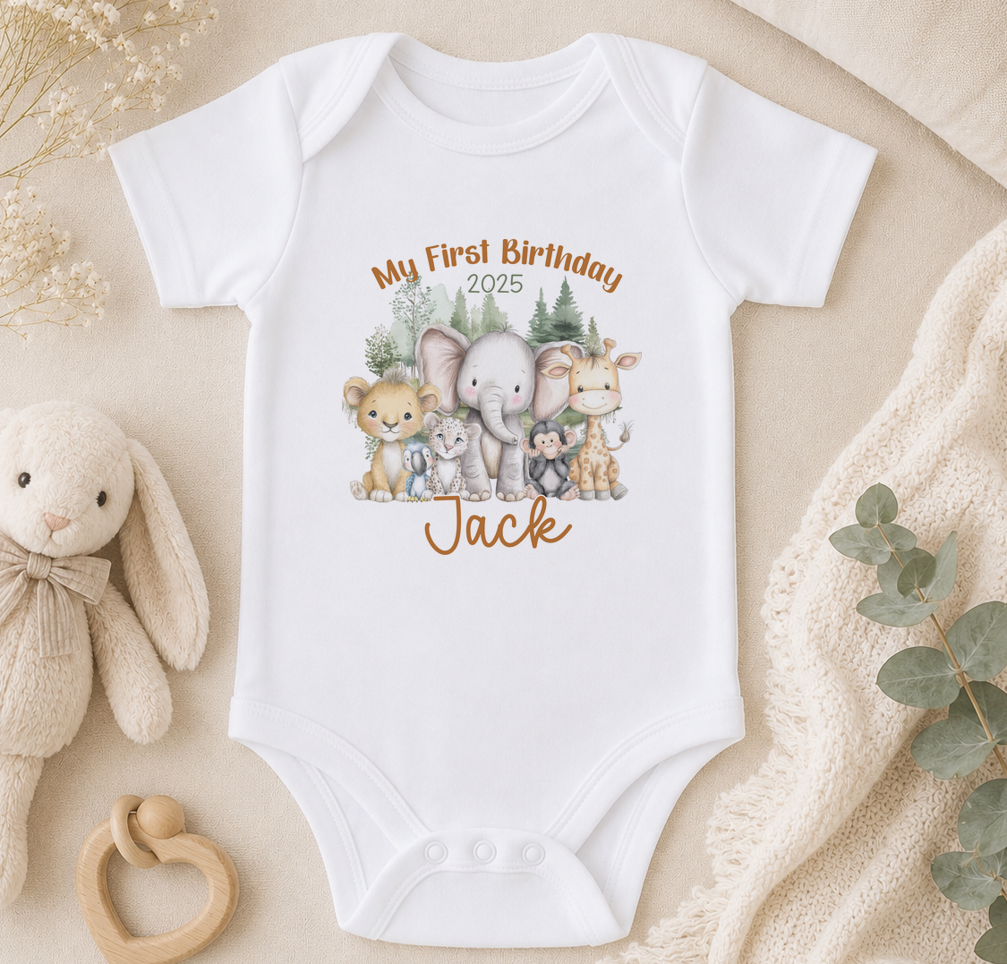 A white baby onesie with a printed image of various animals, including elephants, bunnies, and a giraffe, along with the text "My First Birthday Jack" and the year "2025". The onesie is displayed on a beige surface, surrounded by a stuffed animal, a baby rattle, and some greenery.