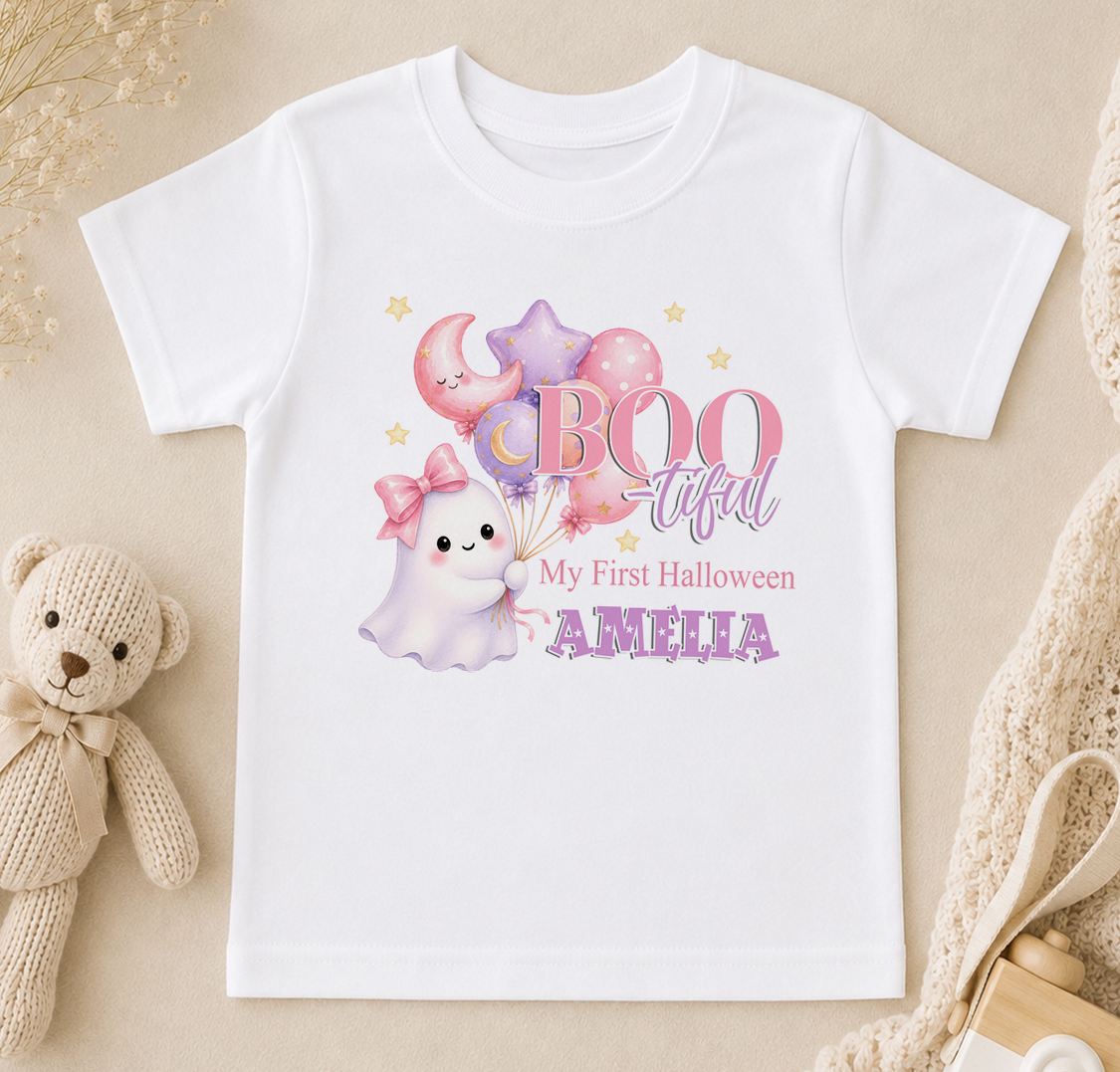 A white t-shirt with a Halloween-themed design featuring a ghost, balloons, and a teddy bear. The text on the t-shirt says "BOO tacular My First Halloween AMELIA".