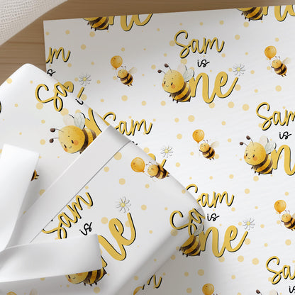 a white gift box with a repeating pattern of yellow bees and the text "Sam is one" printed on it.