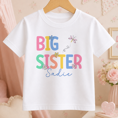 A white t-shirt with the text "BIG SISTER Sadie" printed on it, along with colorful flower designs.