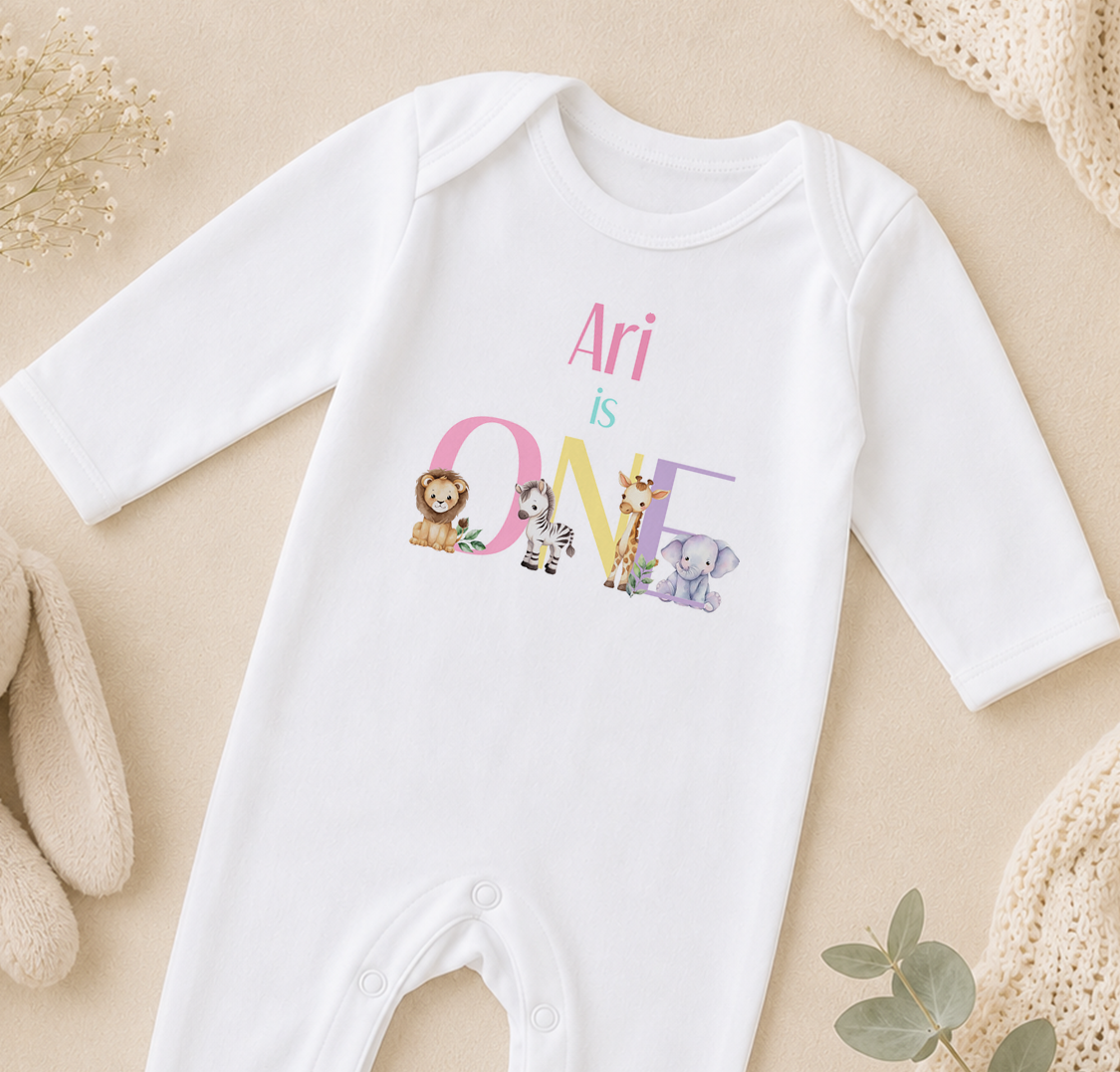 A white onesie with the text "Ari is ONE" printed on it, featuring various animal illustrations.