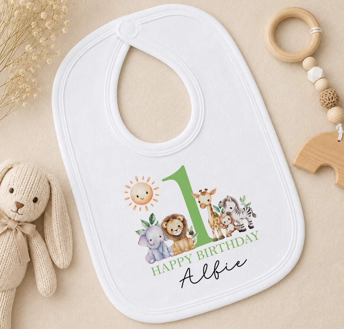 Personalised white bib printed with a green number 1 and animal illustrations and 'Happy Birthday Albie', styled with a soft toy and a wooden bead ring.