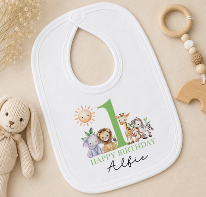 Personalised white bib printed with a green number 1 and animal illustrations and 'Happy Birthday Albie', styled with a soft toy and a wooden bead ring.