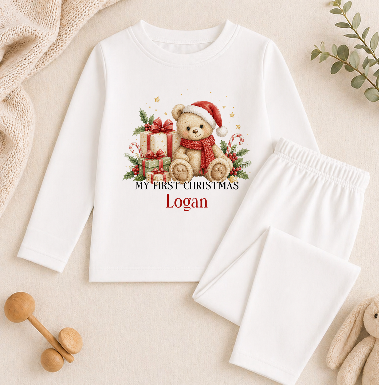 A white long-sleeved shirt with a Christmas-themed graphic of a teddy bear wearing a Santa hat and scarf, along with the text "My first Christmas Logan" printed on it. The shirt is placed on a beige surface, and there are some wooden toys and a plant in the background.