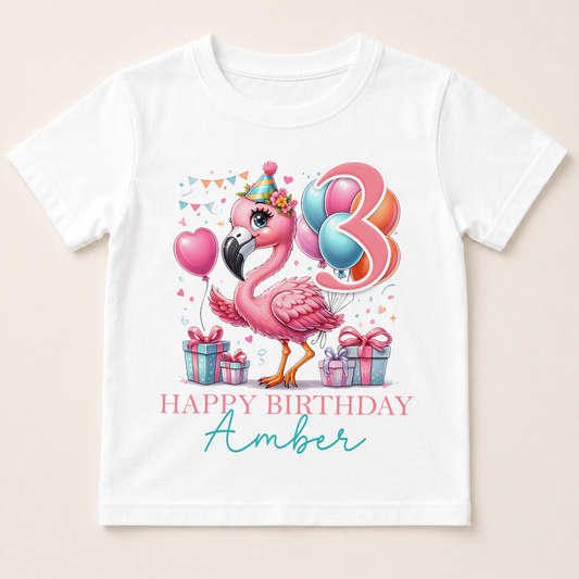 A white t-shirt with a colorful graphic design featuring a pink flamingo surrounded by balloons, presents, and the text "Happy Birthday Amber".