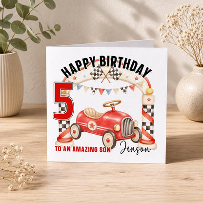 Square birthday card reading Happy Birthday with a red vintage pedal car, chequered racing flags and bunting, personalised To an Amazing Son Jenson 5, standing with its envelope.