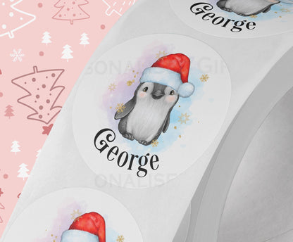 Glossy white round Christmas sticker personalised with a name, printed with a bright colourful party penguin design, shown on a strip of stickers