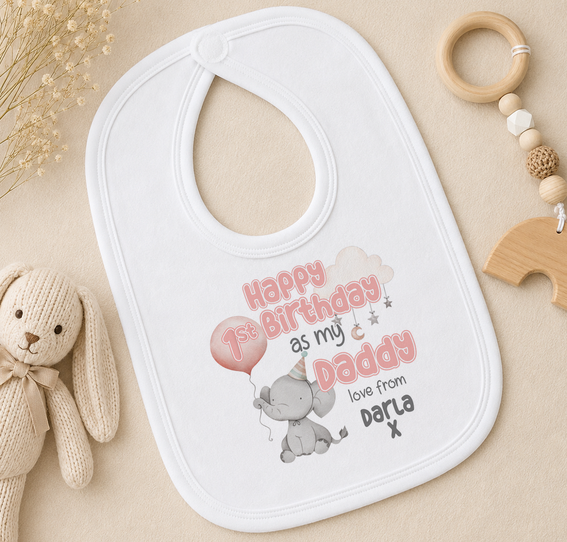 A white baby bib with a personalized message, a wooden toy, and some dried flowers on a beige surface.