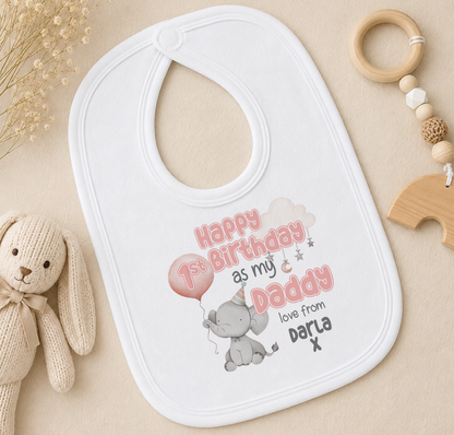 A white baby bib with a personalized message, a wooden toy, and some dried flowers on a beige surface.