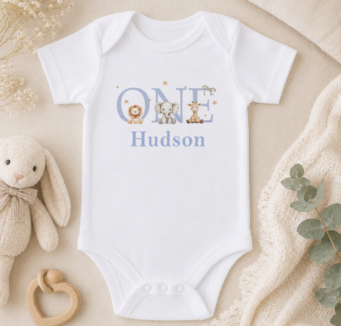 A white onesie with the name "Hudson" printed on it, surrounded by stuffed animals and a beige blanket.