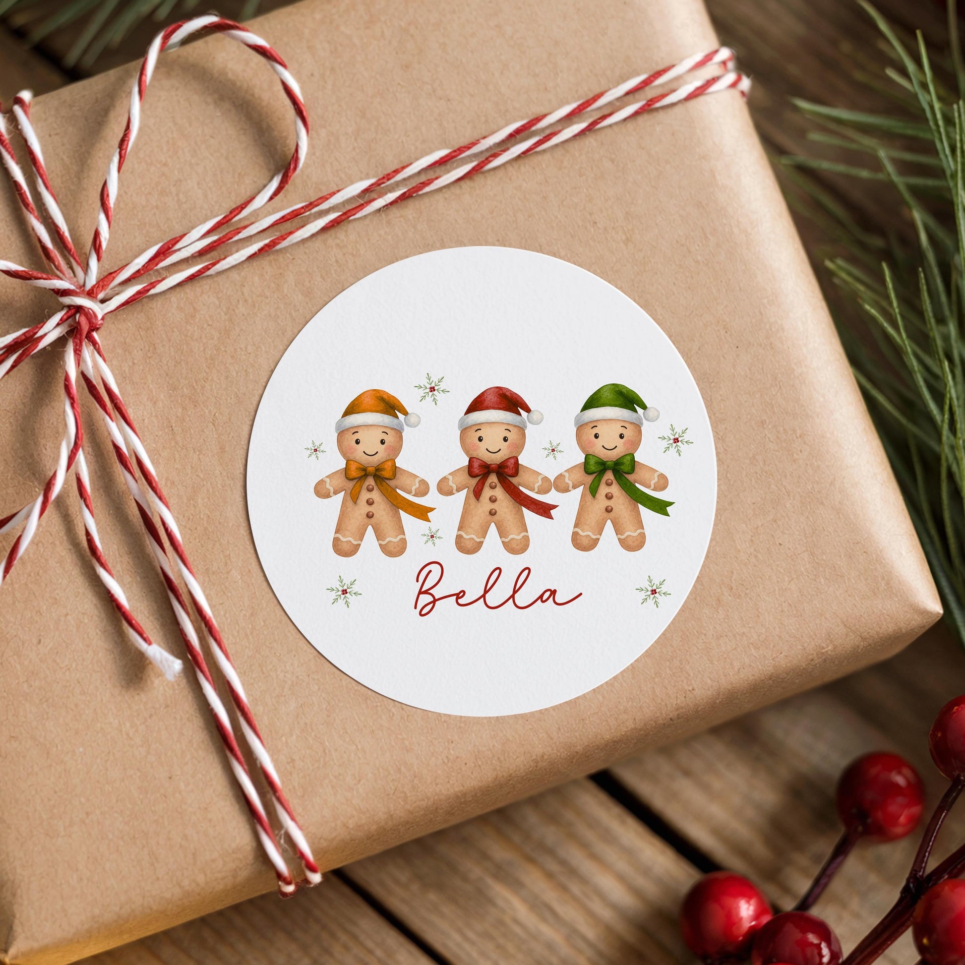 Round white Christmas sticker featuring three smiling gingerbread boys wearing colourful Santa hats with star accents and the personalised name Bella, stuck on a brown gift box tied with red and white twine