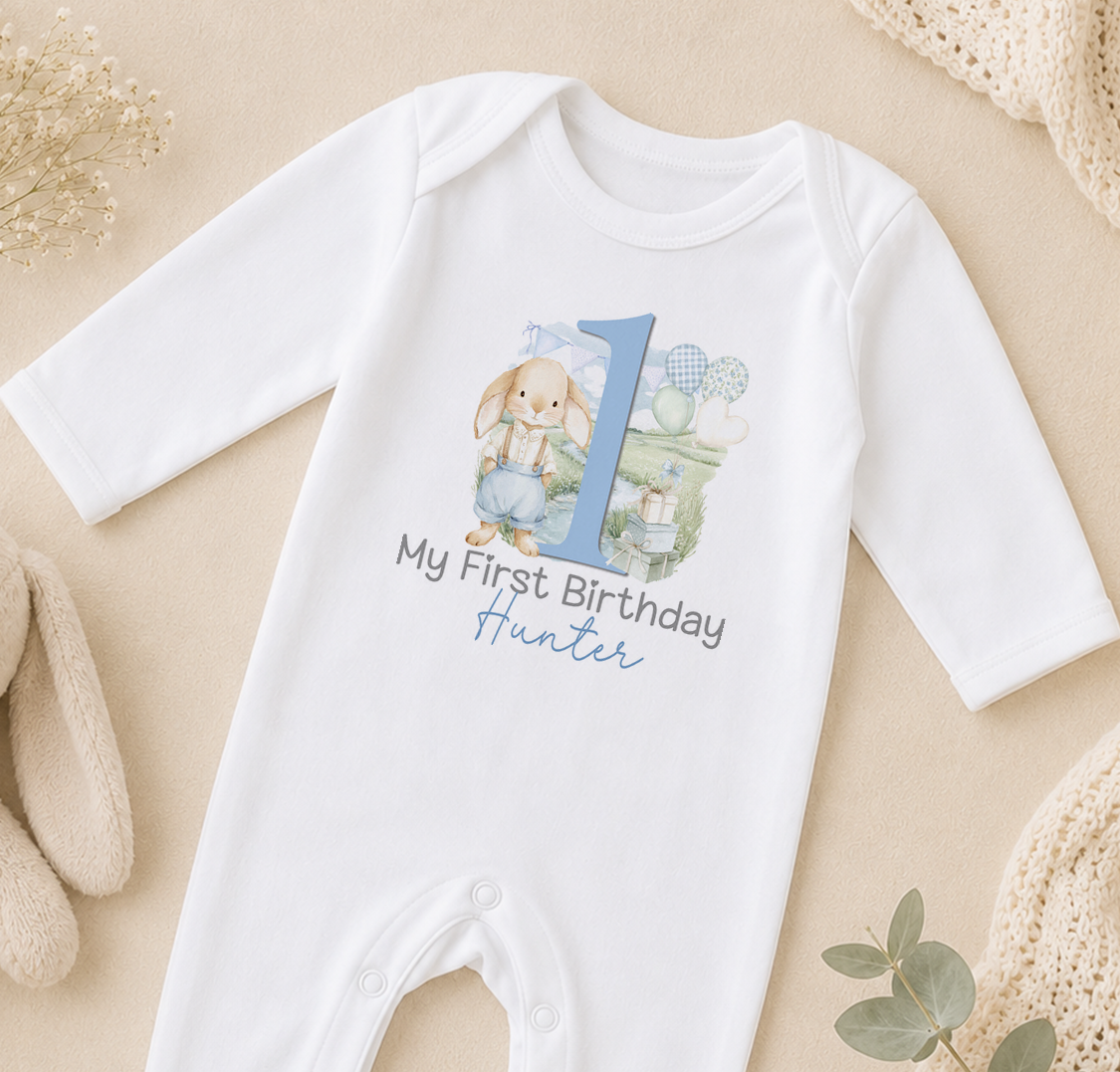 A white onesie with a blue number 1 and the text "My first birthday Hunter" printed on it, along with a bunny illustration.