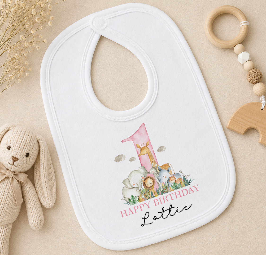 A white baby bib with a pink number one and a giraffe design, placed on a beige surface next to a wooden toy and a wooden bead ring.