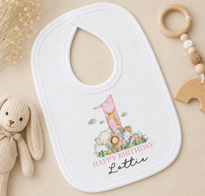 A white baby bib with a pink number one and a giraffe design, placed on a beige surface next to a wooden toy and a wooden bead ring.
