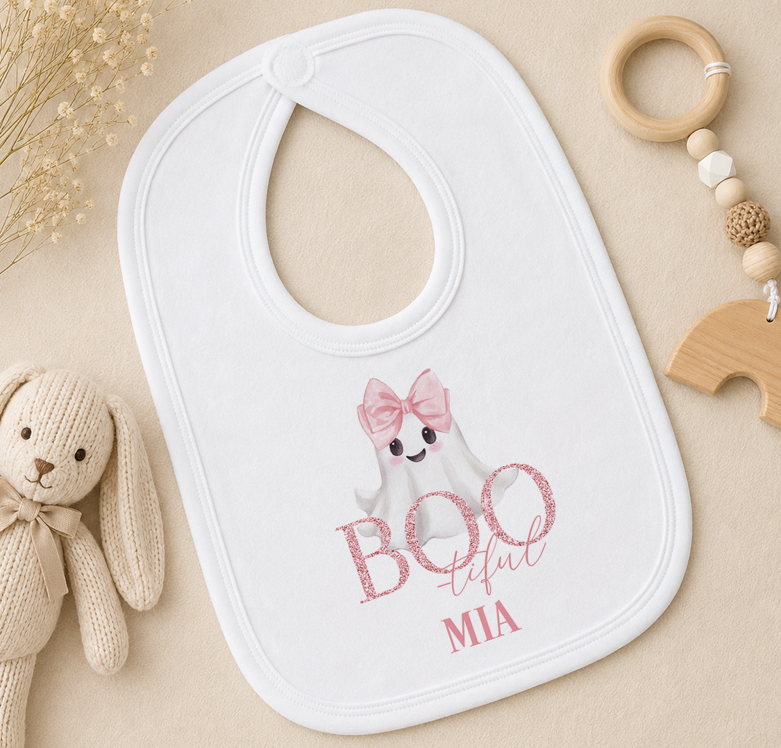 A white baby bib with a ghost character and the text "BOO teful MIA" printed on it, along with a wooden toy and a beaded ringer.