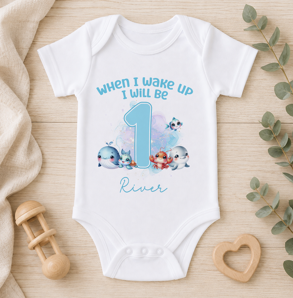 A white onesie with a cartoon image of a baby whale and the text "When I wake up I will be 1 River" printed on it, along with a wooden baby rattle and a wooden heart-shaped object.