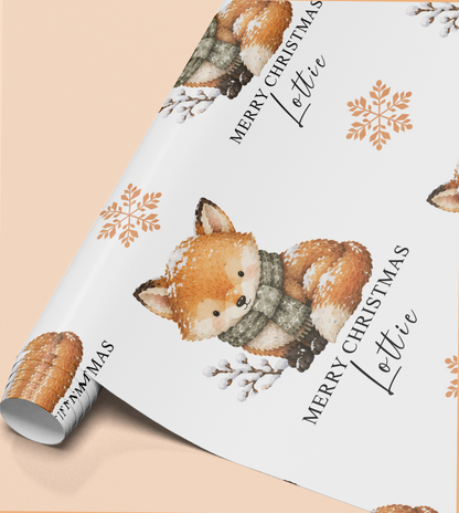 a roll of wrapping paper with a festive design of a fox wearing a scarf and surrounded by snowflakes. The text "Merry Christmas Lottie" is written on the paper, indicating a personalized gift for someone named Lottie.