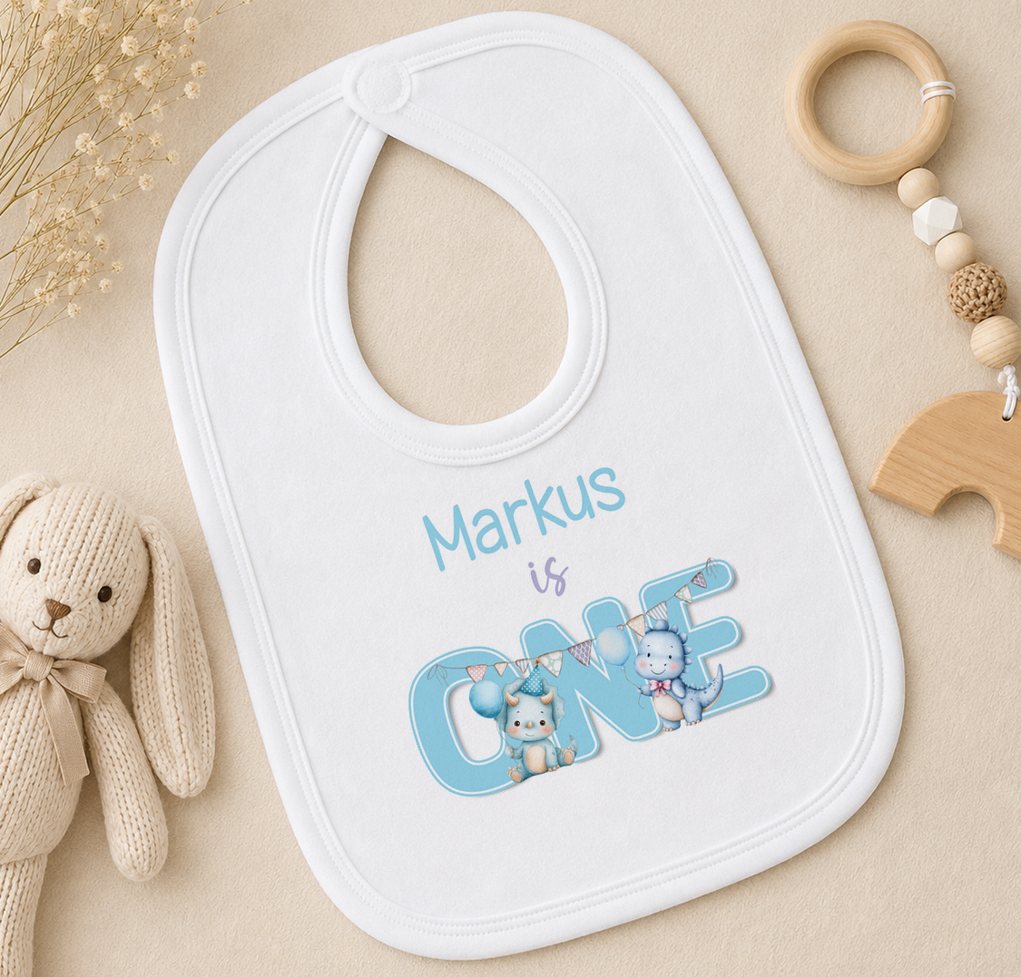 A white bib with the text "Markus is ONE" printed on it, along with illustrations of a bunny and a cat, is placed on a surface next to a wooden toy and a beaded ring.