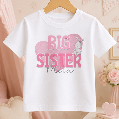 A white t-shirt with a pink graphic and text that says "BIG SISTER Mila" hanging on a wooden hanger.