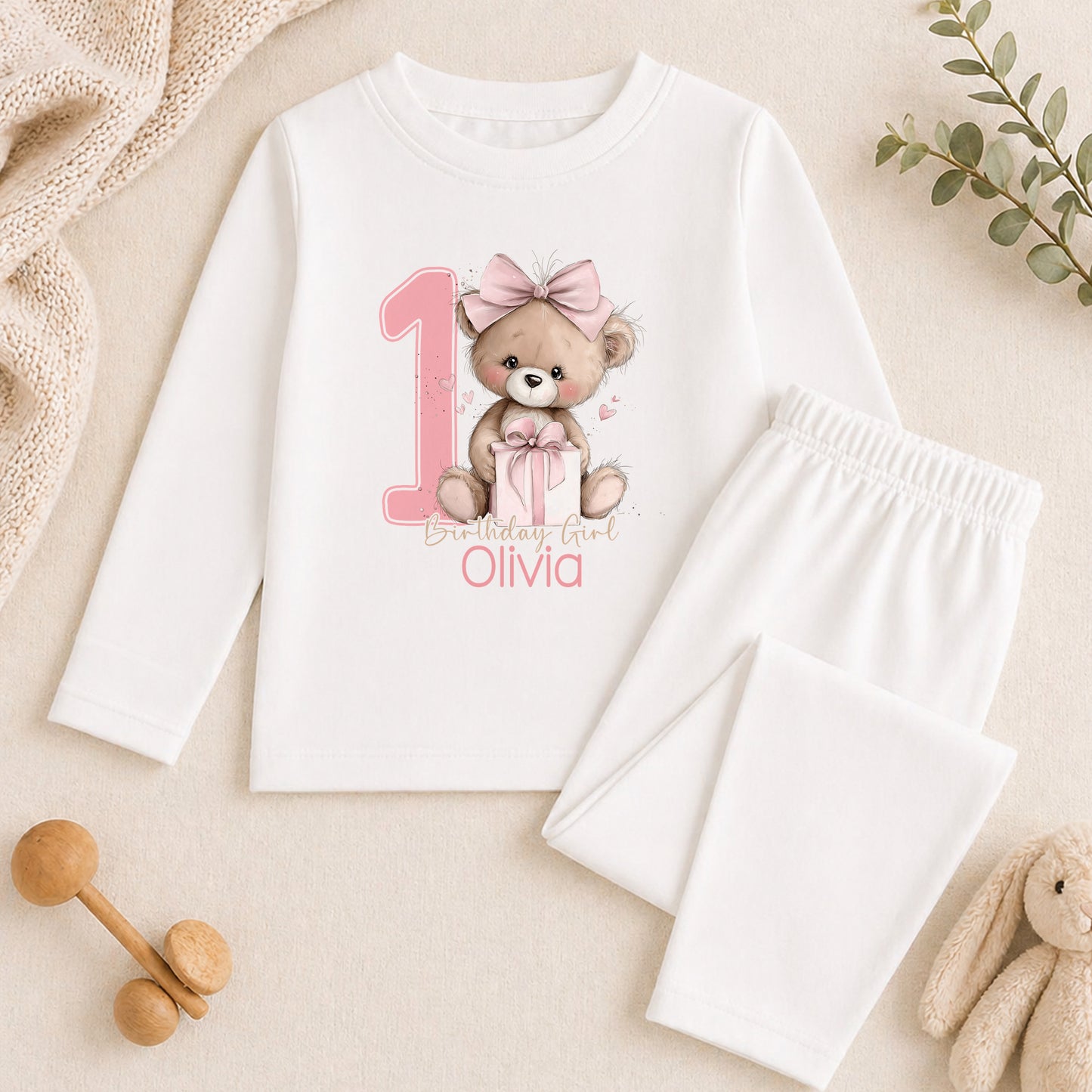 A white long-sleeved shirt with a teddy bear graphic and the number 1, along with a pair of white pajama pants, are displayed on a beige surface. There are also some stuffed animals and a plant in the background.