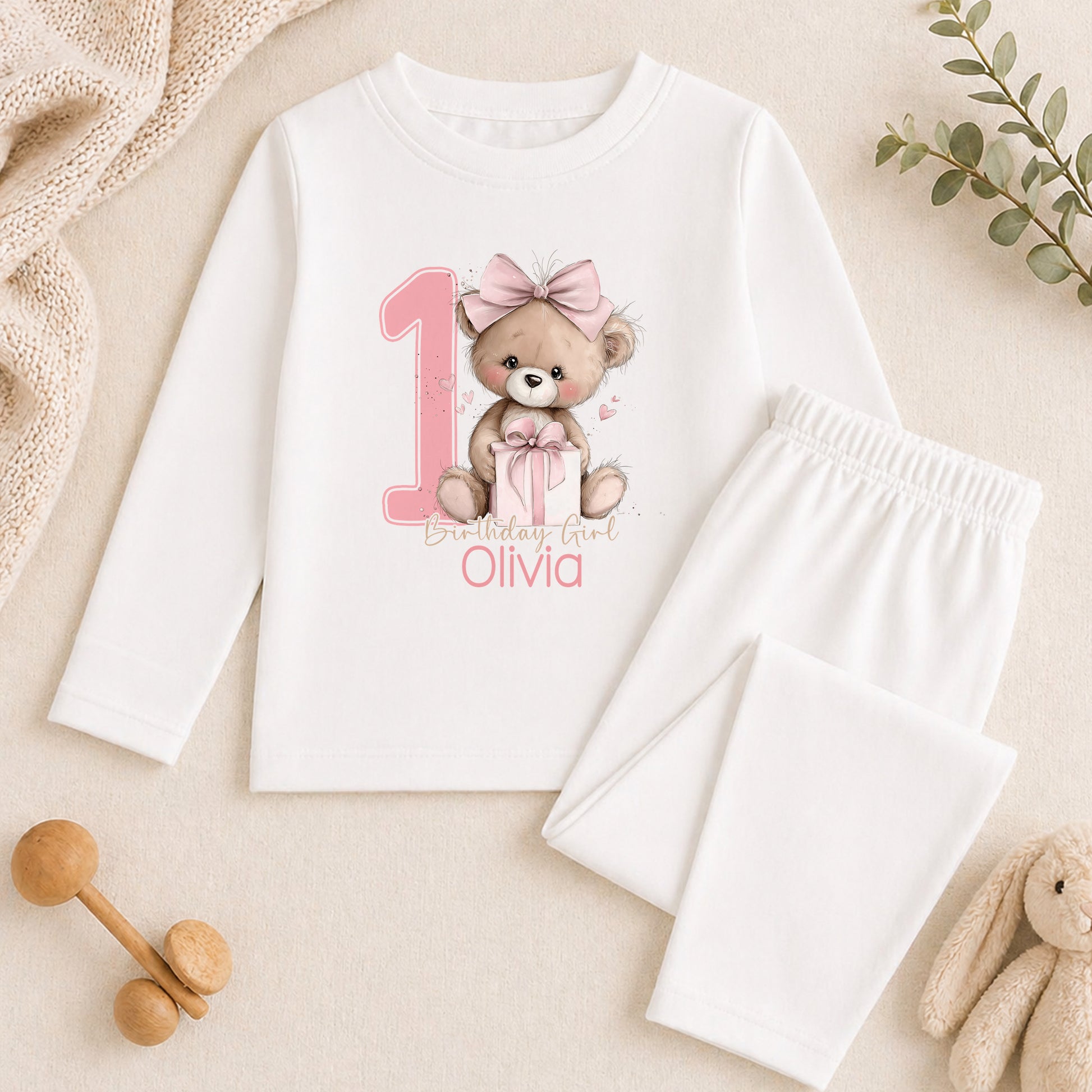 A white long-sleeved shirt with a teddy bear graphic and the number 1, along with a pair of white pajama pants, are displayed on a beige surface. There are also some stuffed animals and a plant in the background.