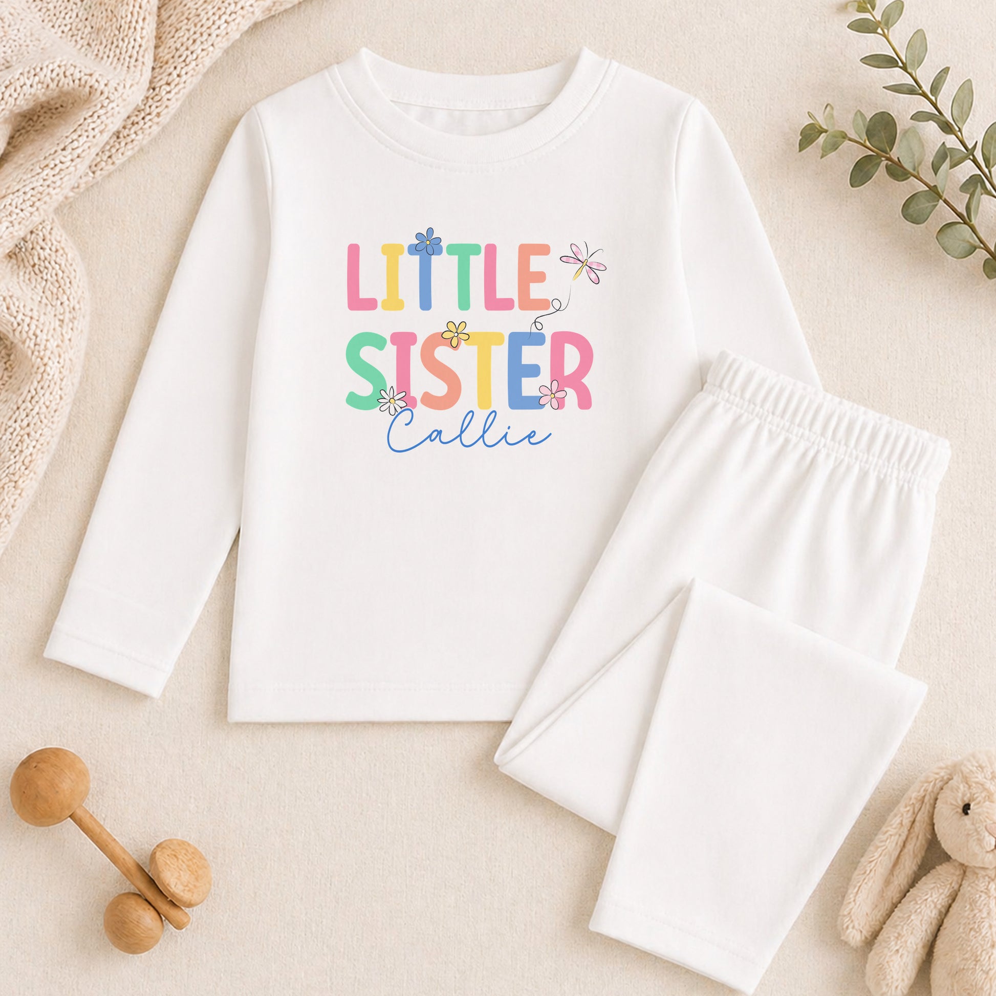 A white long-sleeved shirt with the text "Little Sister Callie" printed on it, along with a pair of white pajama pants. The shirt and pants are placed on a beige surface, and there are some toys and greenery in the background.