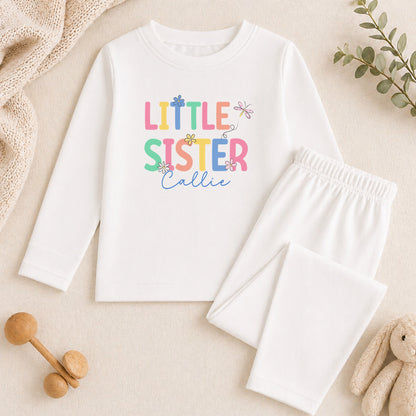 A white long-sleeved shirt with the text "Little Sister Callie" printed on it, along with a pair of white pajama pants. The shirt and pants are placed on a beige surface, and there are some toys and greenery in the background.