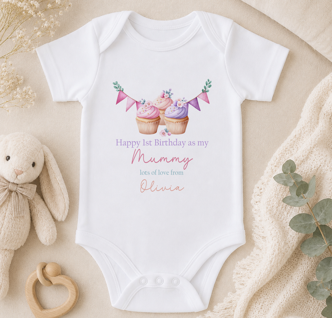 A white onesie with a birthday message for a baby, featuring cupcakes and a ribbon, placed on a beige surface with a stuffed animal and greenery.