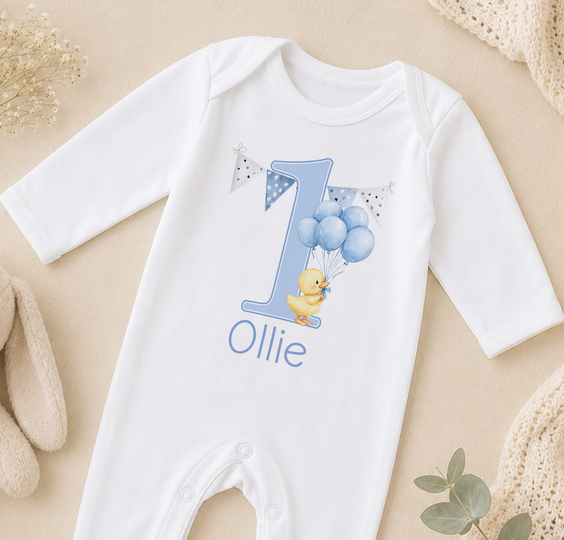 A white onesie with a blue number one and a yellow duck wearing balloons, along with the name "Ollie" printed on it.
