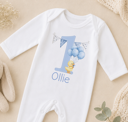 A white onesie with a blue number one and a yellow duck wearing balloons, along with the name "Ollie" printed on it.