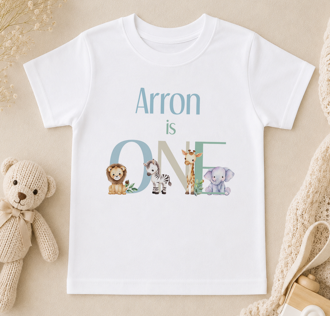 A white t-shirt with the text "Arron is ONE" printed on it, along with illustrations of various animals such as a lion, zebra, giraffe, and elephant.