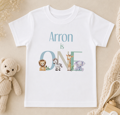 A white t-shirt with the text "Arron is ONE" printed on it, along with illustrations of various animals such as a lion, zebra, giraffe, and elephant.