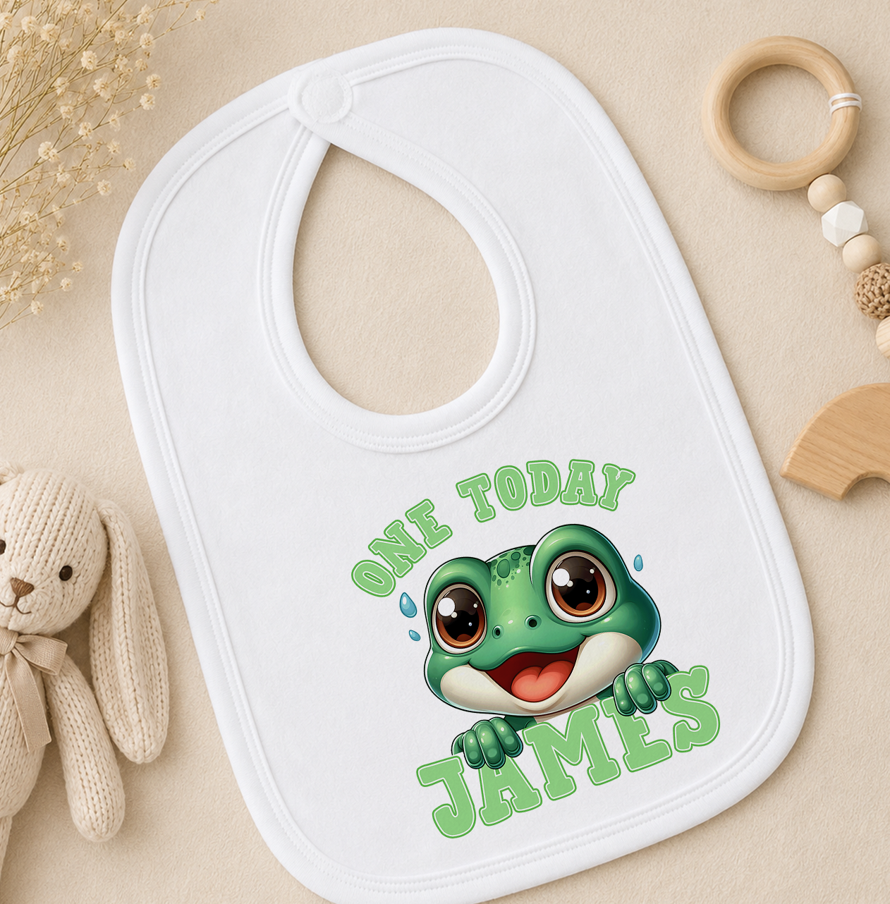 White personalised baby bib printed with a cartoon frog and 'One Today James', shown with a wooden toy and beaded rattle.