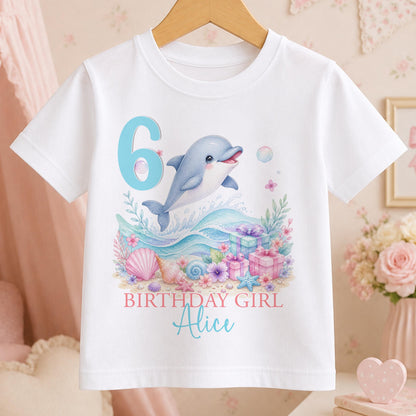 White cotton t-shirt printed with a watercolour dolphin leaping over pastel waves beside a large age number, with Birthday Girl and a name below.