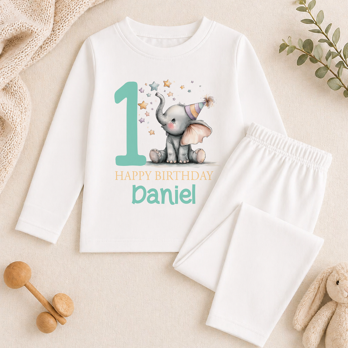 A white long-sleeved shirt with a graphic of an elephant and the text "Happy Birthday Daniel" printed on it, along with a pair of white pajama pants. The shirt and pants are placed on a beige surface, and there are some toys and greenery in the background.