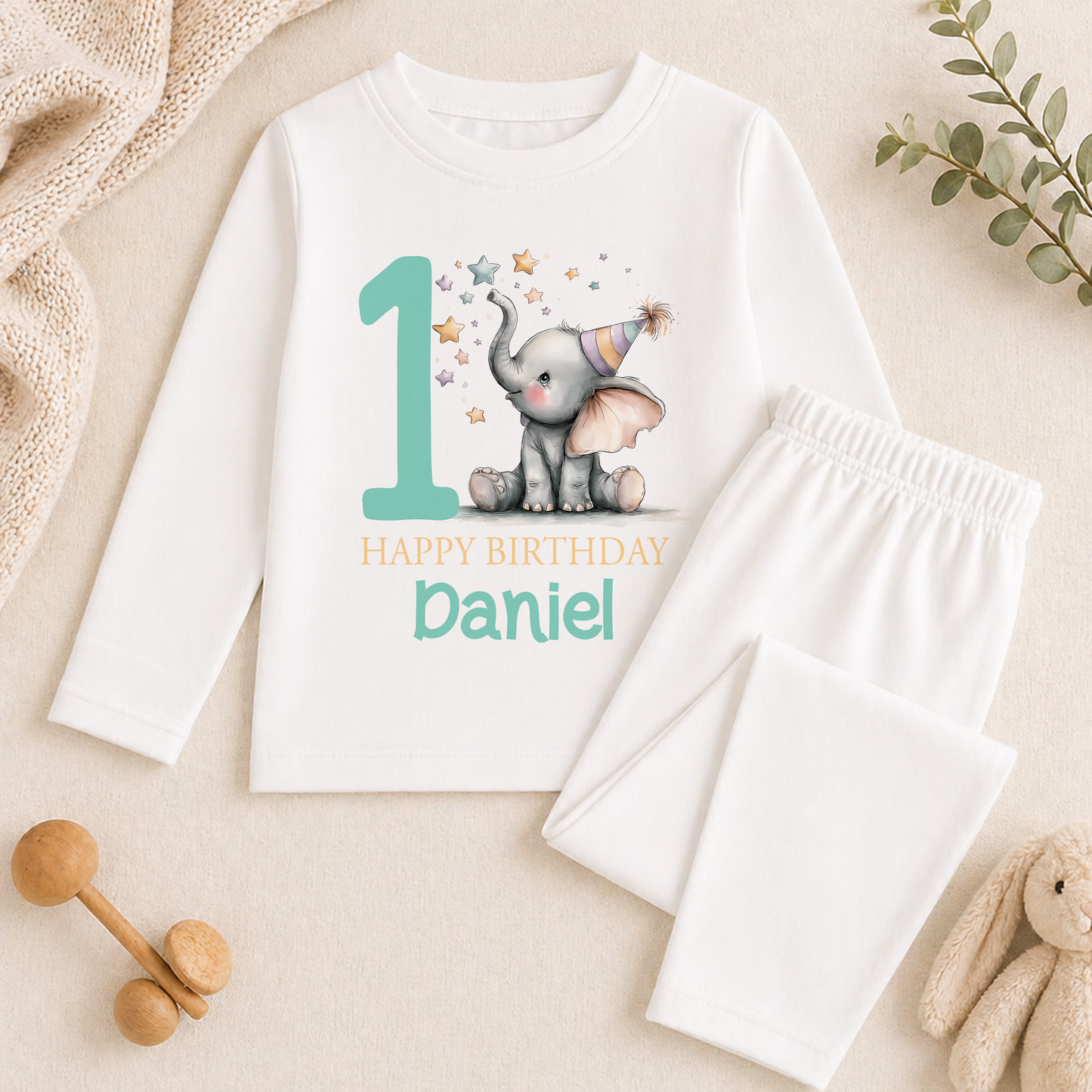 A white long-sleeved shirt with a graphic of an elephant and the text "Happy Birthday Daniel" printed on it, along with a pair of white pajama pants. The shirt and pants are placed on a beige surface, and there are some toys and greenery in the background.