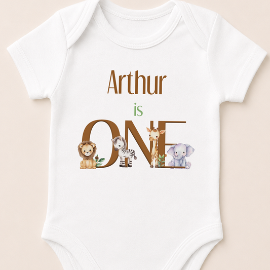 A white baby onesie with the text "Arthur is ONE" and illustrations of various animals, including a lion, zebra, giraffe, and elephant.