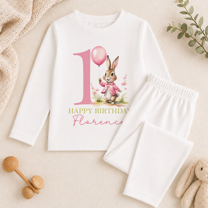 A white long-sleeved shirt with a pink bunny and a balloon, along with a pair of white pants, are displayed on a beige surface. There are also some stuffed animals and a plant in the background.
