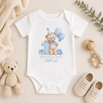 Personalised first birthday outfit in white with a teddy bear in a blue party hat, blue balloons and presents, Birthday Boy with a name and age