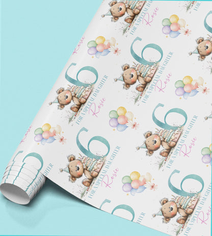 a roll of wrapping paper with a repeating pattern of a teddy bear wearing a party hat and holding a birthday cake, along with colorful balloons.