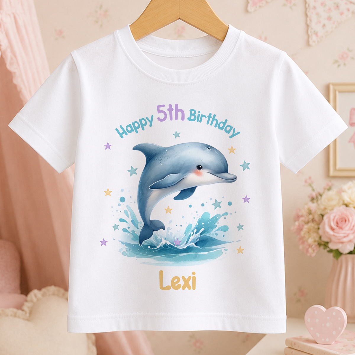 A white t-shirt with a cartoon image of a dolphin jumping out of the water, surrounded by stars and a pink background. The text on the t-shirt reads "Happy 5th Birthday Lexi".