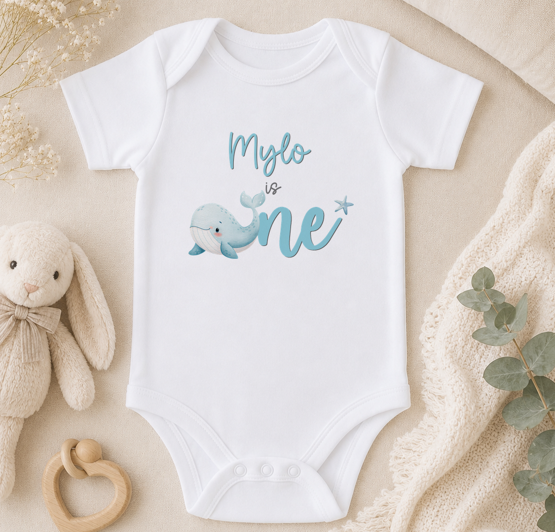 A white onesie with a blue whale graphic and the text "Mylo is one" is displayed on a beige surface, accompanied by a stuffed animal and a plant.