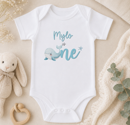 A white onesie with a blue whale graphic and the text "Mylo is one" is displayed on a beige surface, accompanied by a stuffed animal and a plant.