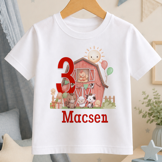 White T-shirt printed with a red barn, farmyard animals, the number 3 and the personalised name Macsen, displayed on a hanger.