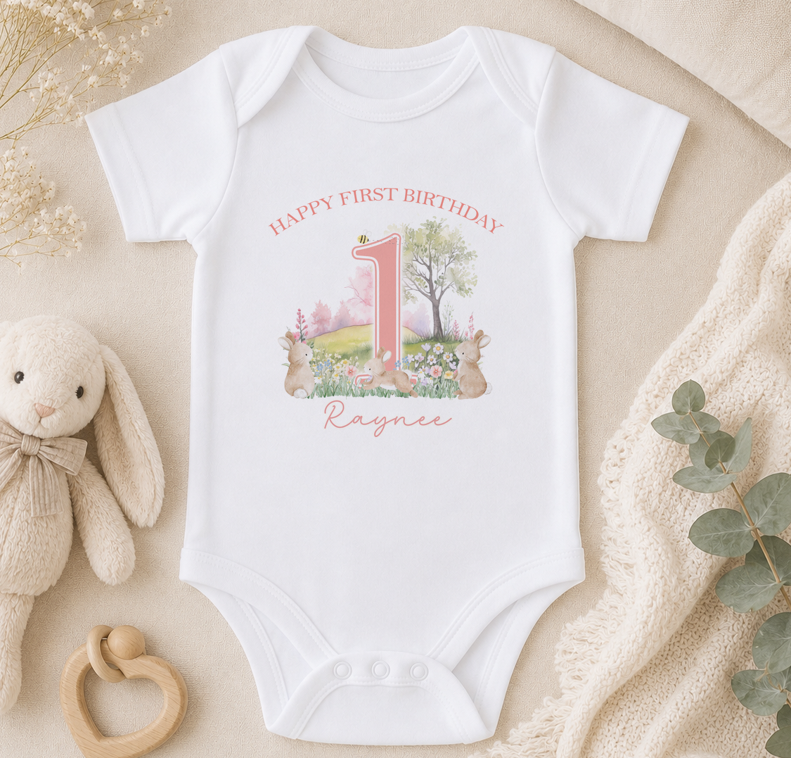 A white onesie with a pink number 1 and a bunny illustration, along with a stuffed bunny and a beige blanket, are arranged on a surface.