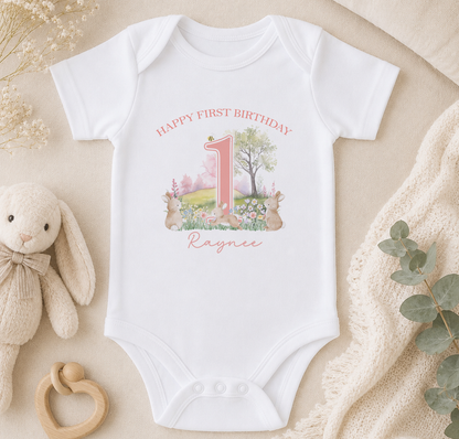 A white onesie with a pink number 1 and a bunny illustration, along with a stuffed bunny and a beige blanket, are arranged on a surface.