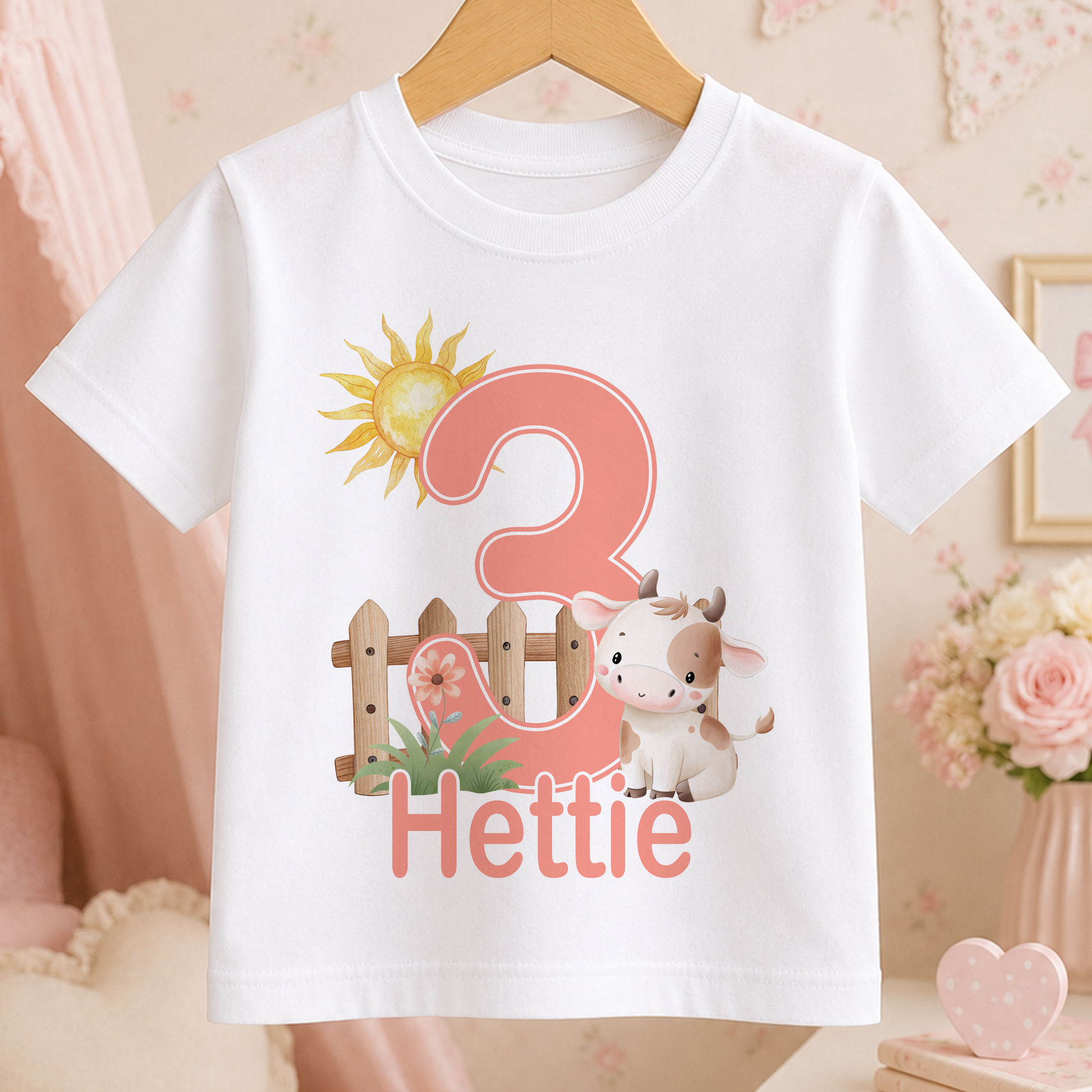A white t-shirt with a pink number 3 and a cartoon cow printed on it, along with the text "Hettie" below the number.