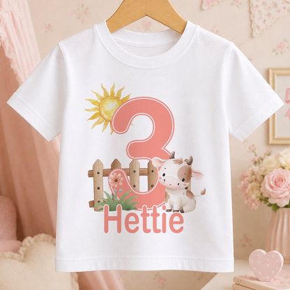 A white t-shirt with a pink number 3 and a cartoon cow printed on it, along with the text "Hettie" below the number.